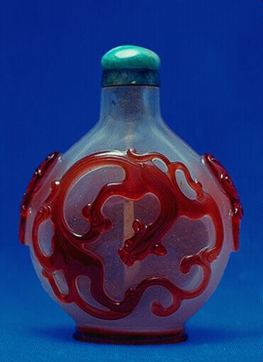 Glass, the milky translucent snuff bottle with a ruby red overlay, carved on each side with a curled, writhing chilong, its tail and limbs creating a circular pattern, the shoulders with unusual mask and ring handles derived from ancient Chinese bronzes.