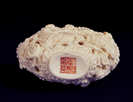 The base with an iron red Jiaqing mark of the porcelain snuff bottle, creamy-white enamel; moulded and reticulated with the design of Fo lions chasing beribboned brocade balls; set against a wave design the base and shoulders surrounded by ruyi lappets and the neck with a key-fret border.