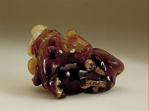 A rich dark brown chalcedony stone, with pale beige and almost white inclusions, brilliantly carved as a reclining horse with a couple of monkeys. One monkey is white, the other beige, and the horse is dark brown.