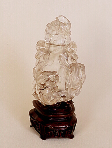 A clear rock crystal Vase, carved with lingzhi, peach, and a flower plus a sack. A duck, with a peach branch in its beak, forms the cover, with loose ring handles either side.